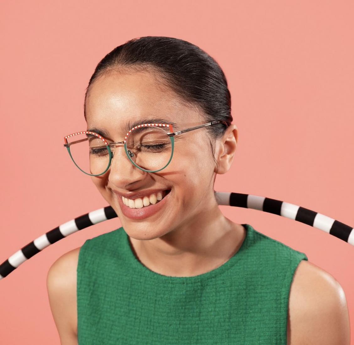 A young woman wearing round glasses and a green top, smiling broadly against a pink background
