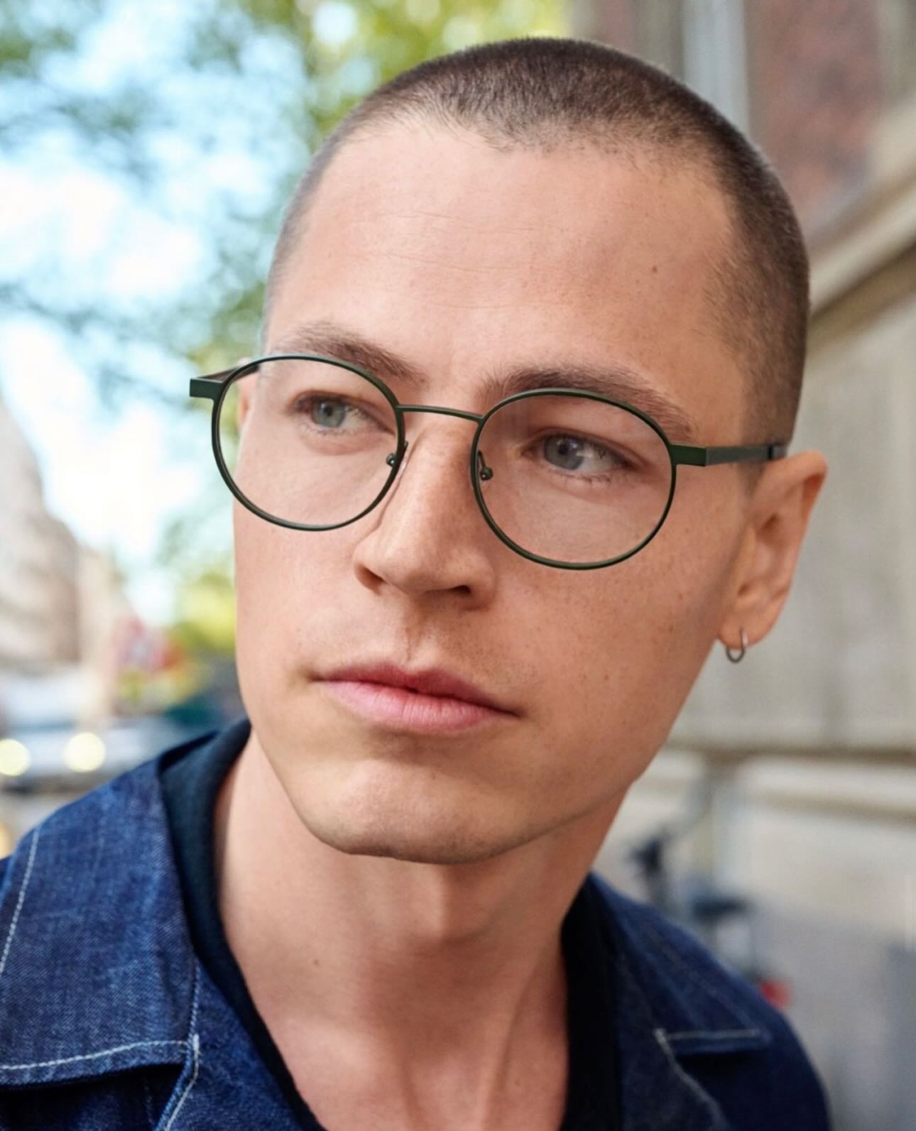 A young man with a shaved head wearing round wire-framed glasses, photographed outdoors in natural light
