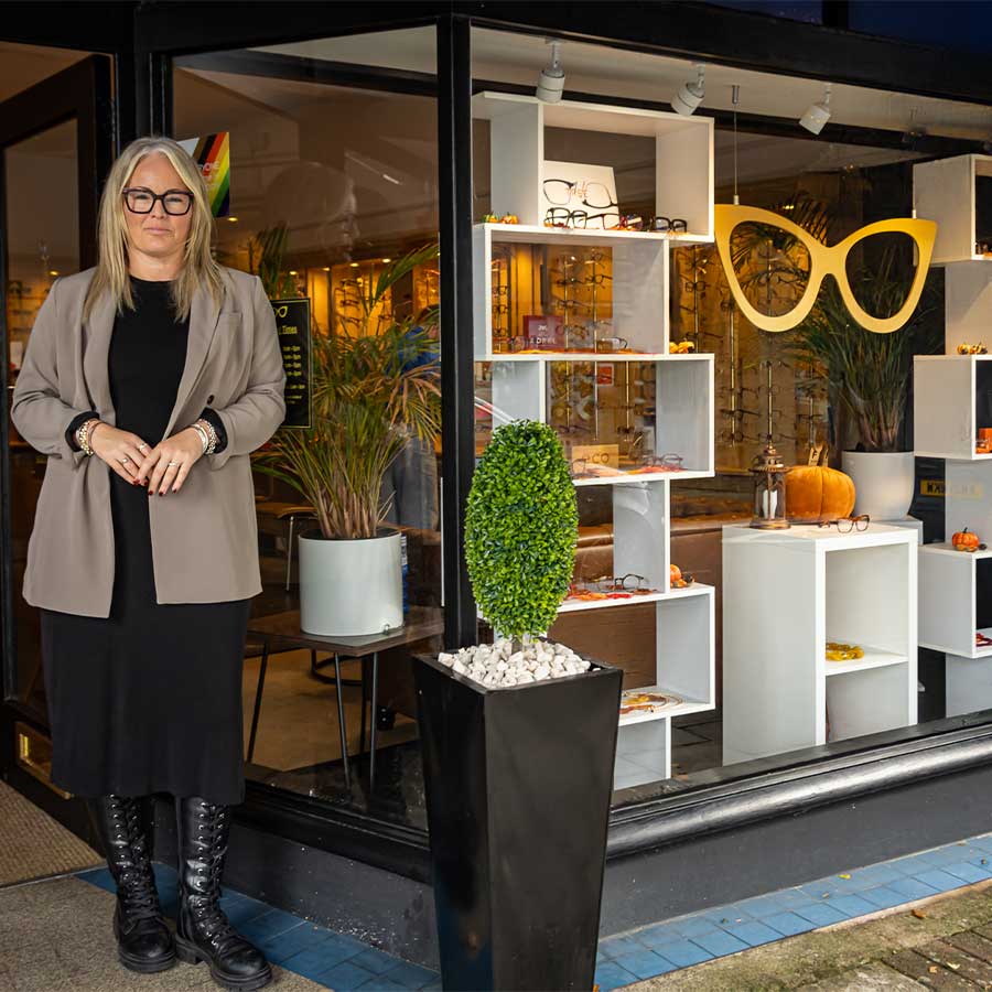 A woman with blonde hair wearing glasses and a grey blazer, standing outside the Tudball Opticians shopfront with the practice's distinctive window display visible behind her