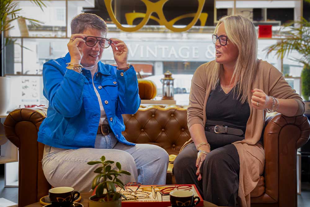 A women in a blue jacket and a woman in a beige cardigan sitting together on a leather sofa, both trying on glasses frames inside the optical practice