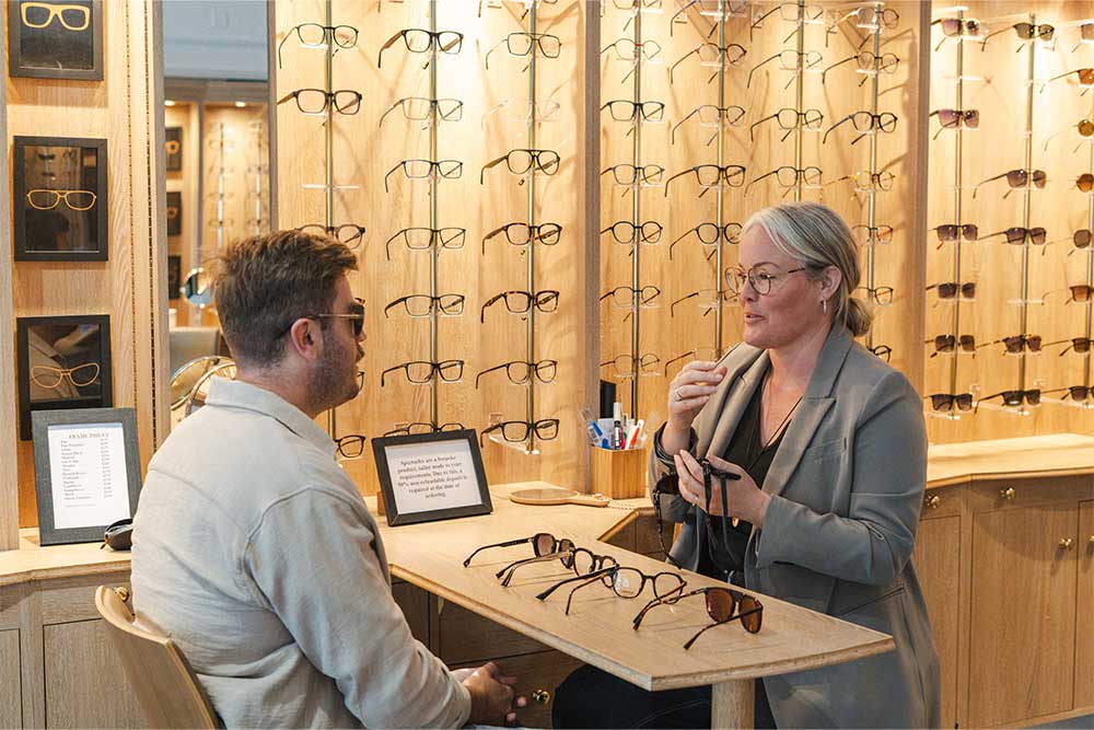 A dispensing optician sitting across from a customer at a desk during an eyewear consultation, with a wall of glasses frames visible behind them