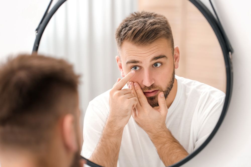 A man inserting a contact lens while looking in a bathroom mirror