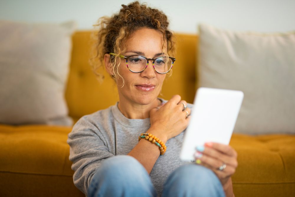 A woman with curly hair wearing glasses, sitting on a yellow sofa and holding a tablet, smiling warmly