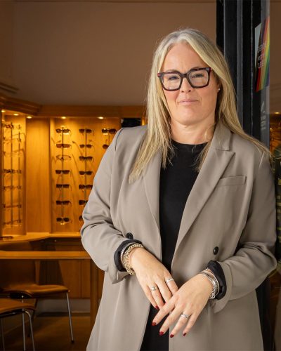 A woman with blonde hair wearing rectangular glasses, dressed in a grey blazer, standing in front of a wall of eyewear displays