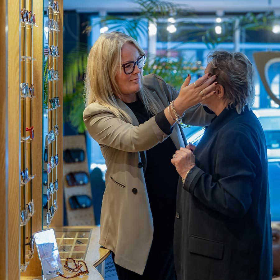 An optician helping a female customer try on glasses inside the Tudball Opticians practice, with eyewear displays visible in the background