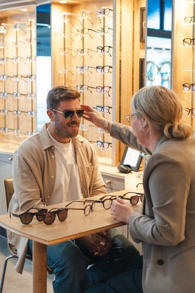 An optician sitting alongside a customer during a relaxed eyewear consultation, with glasses frames visible on the table between them