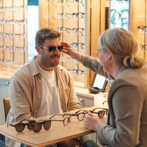 An optician sitting alongside a customer during a relaxed eyewear consultation, with glasses frames visible on the table between them