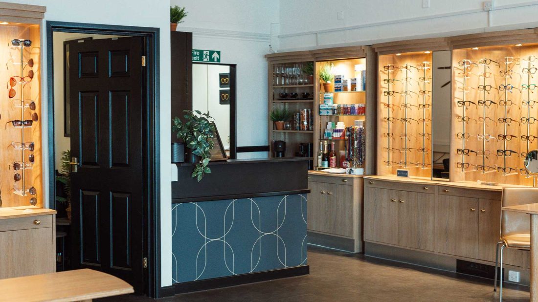 The welcoming interior of Tudball Opticians showing a reception desk, warm wood panelling, and illuminated wall displays of eyewear frames